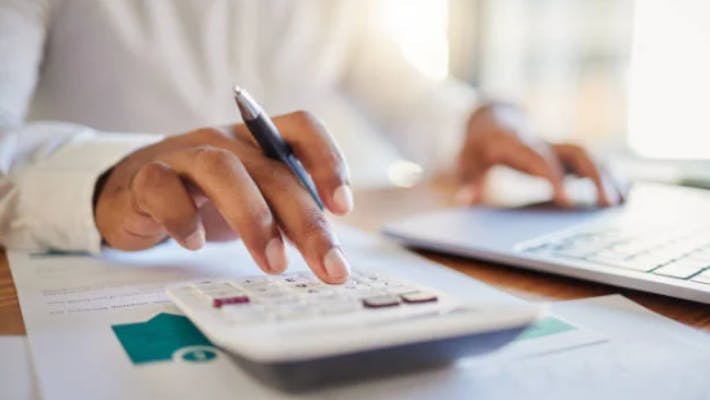 Person with pen in hand using laptop and calculator with paperwork beside