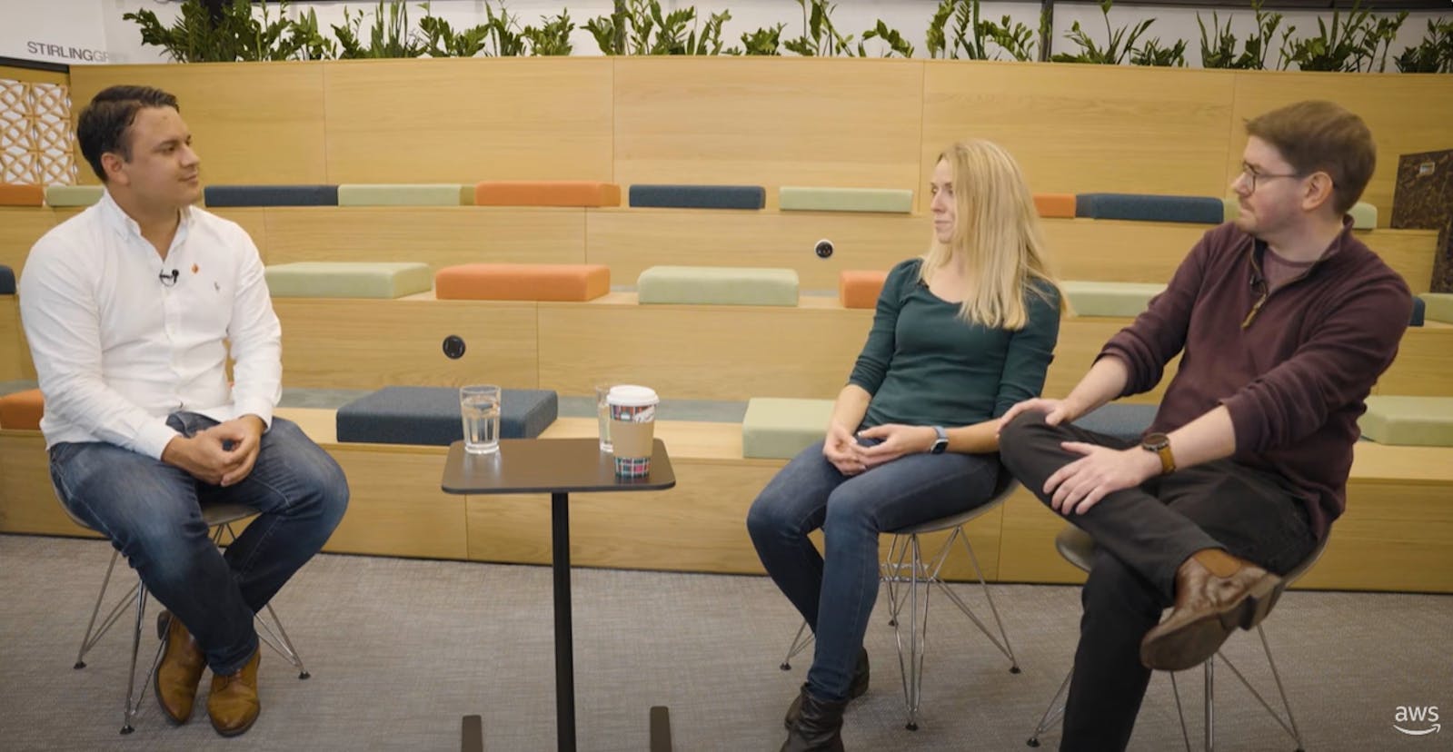 Studio interview with Mo Das from AWS and Lucy Rickards and Richard Huston from Alfa. Stepped wooden seating with colourful green and orange seat cushions in the background